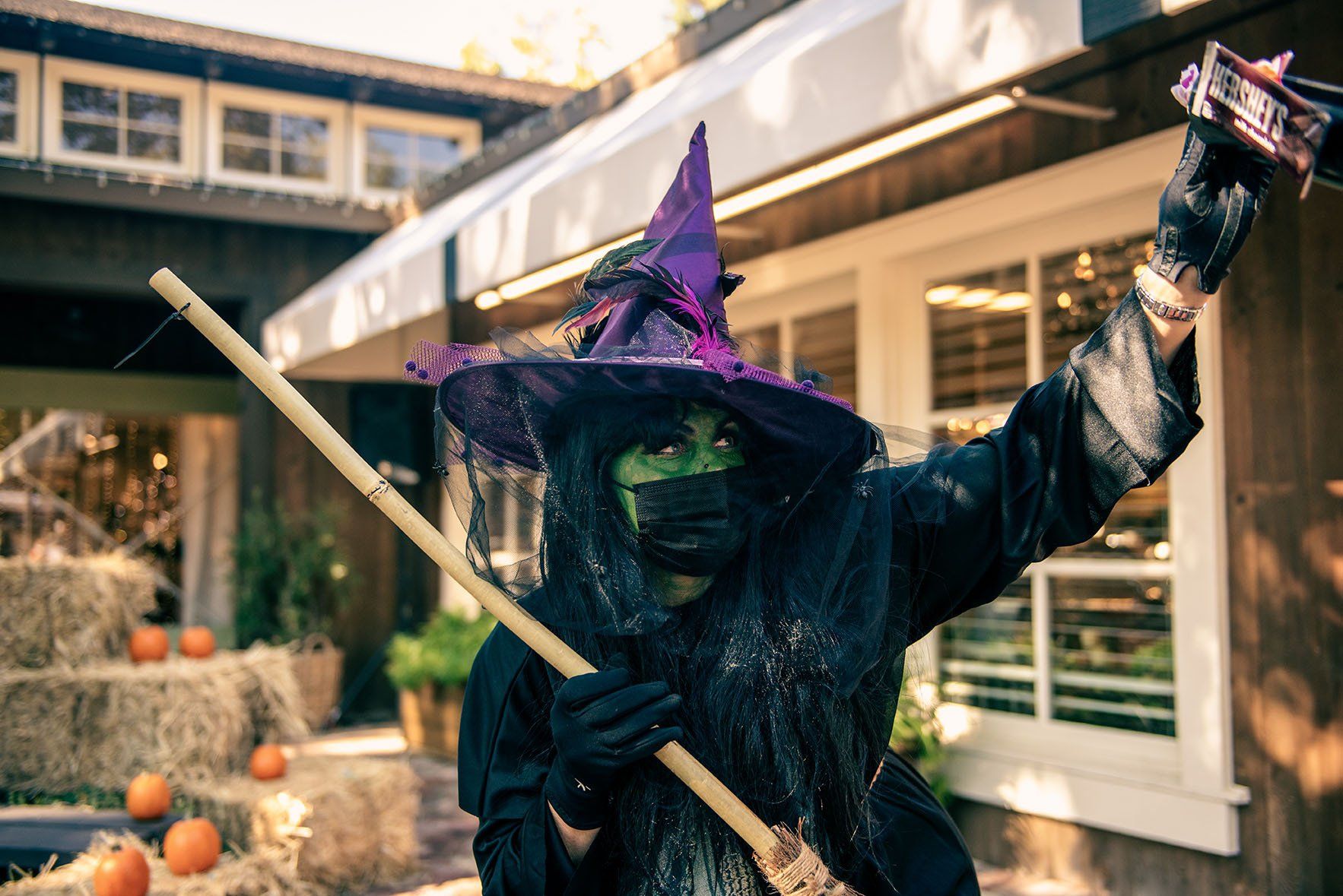 A person dressed as a witch is holding a broom in front of a house.