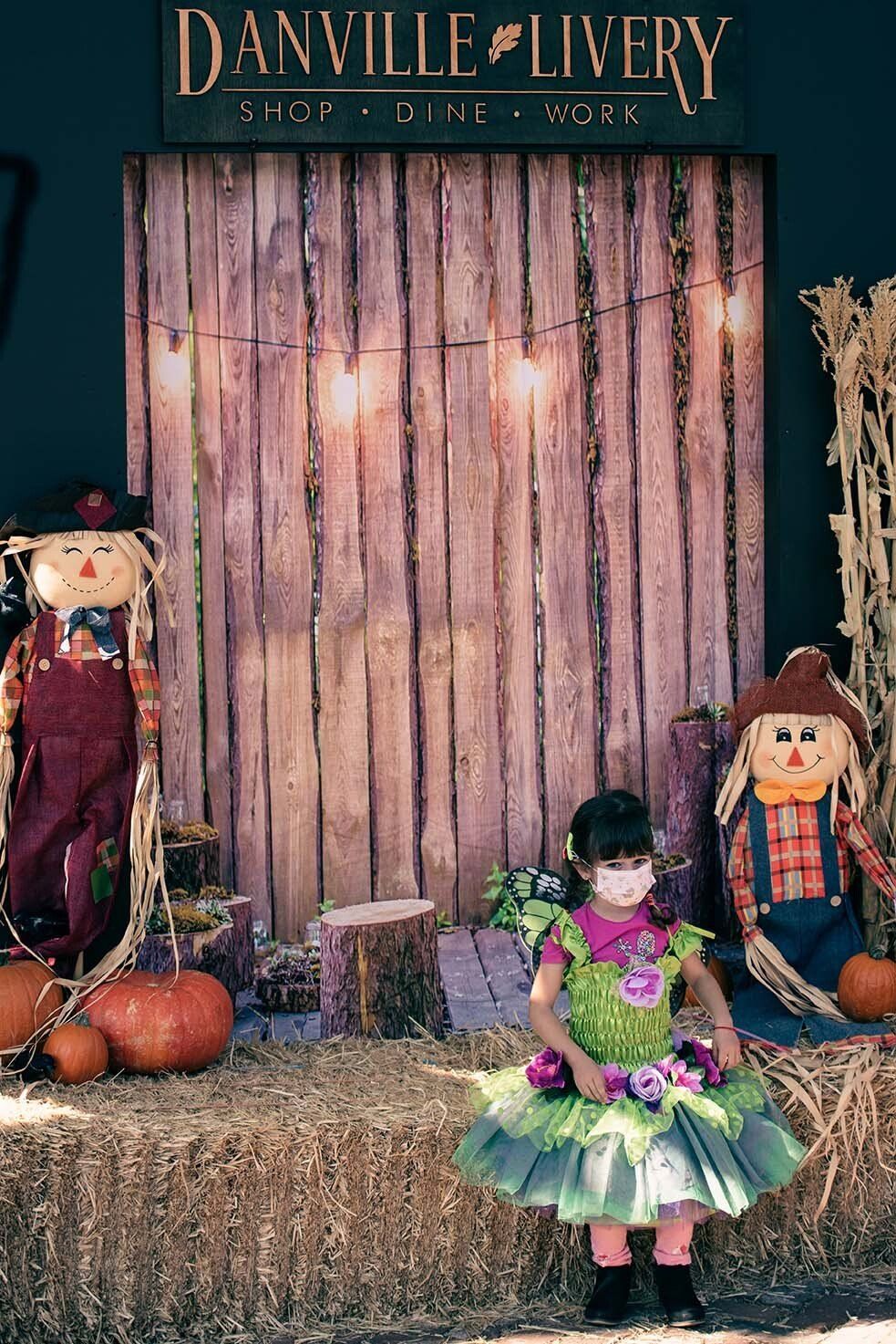 A little girl wearing a mask is standing in front of a wooden wall.