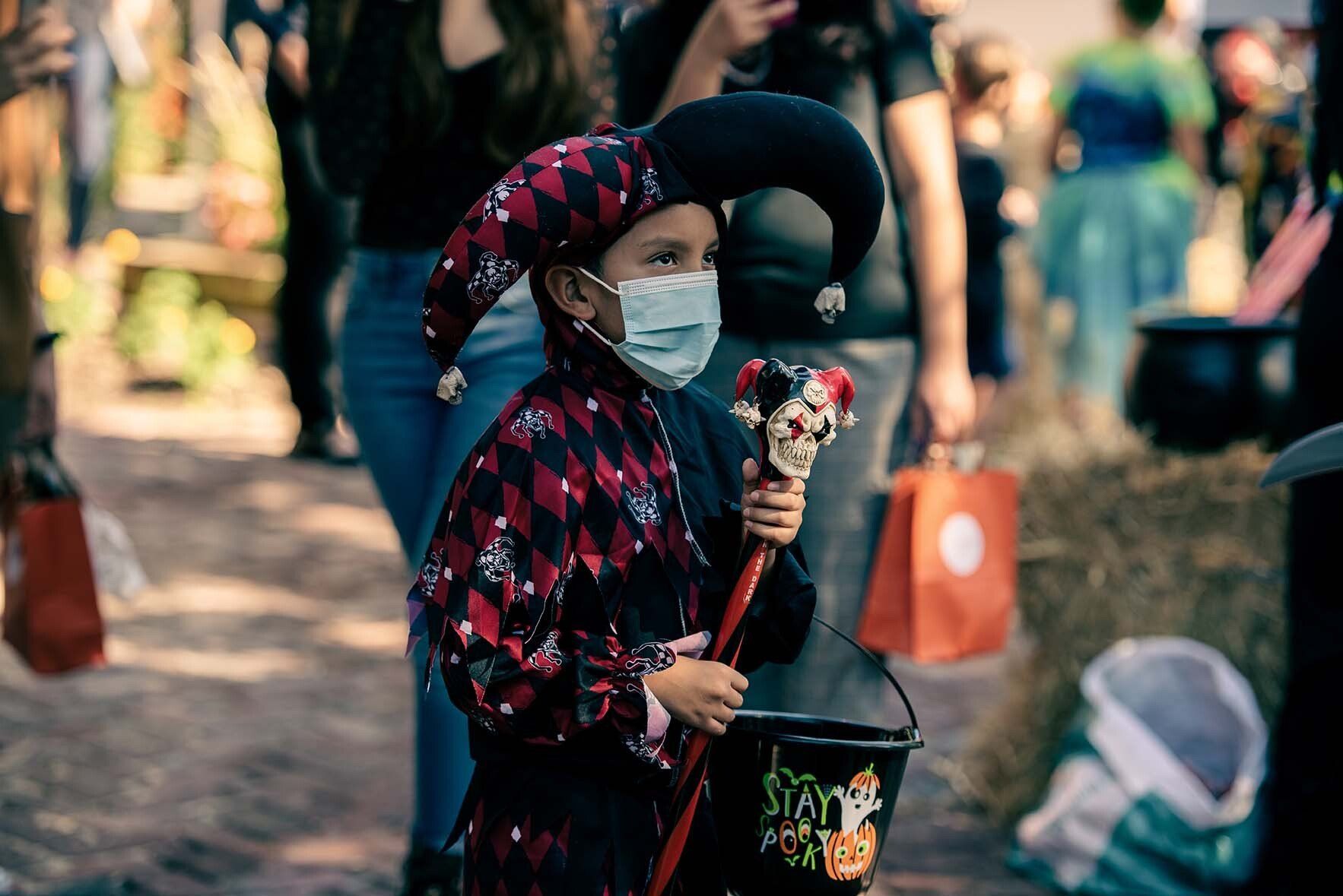A young boy wearing a mask and a jester costume is holding a trick or treat bucket.