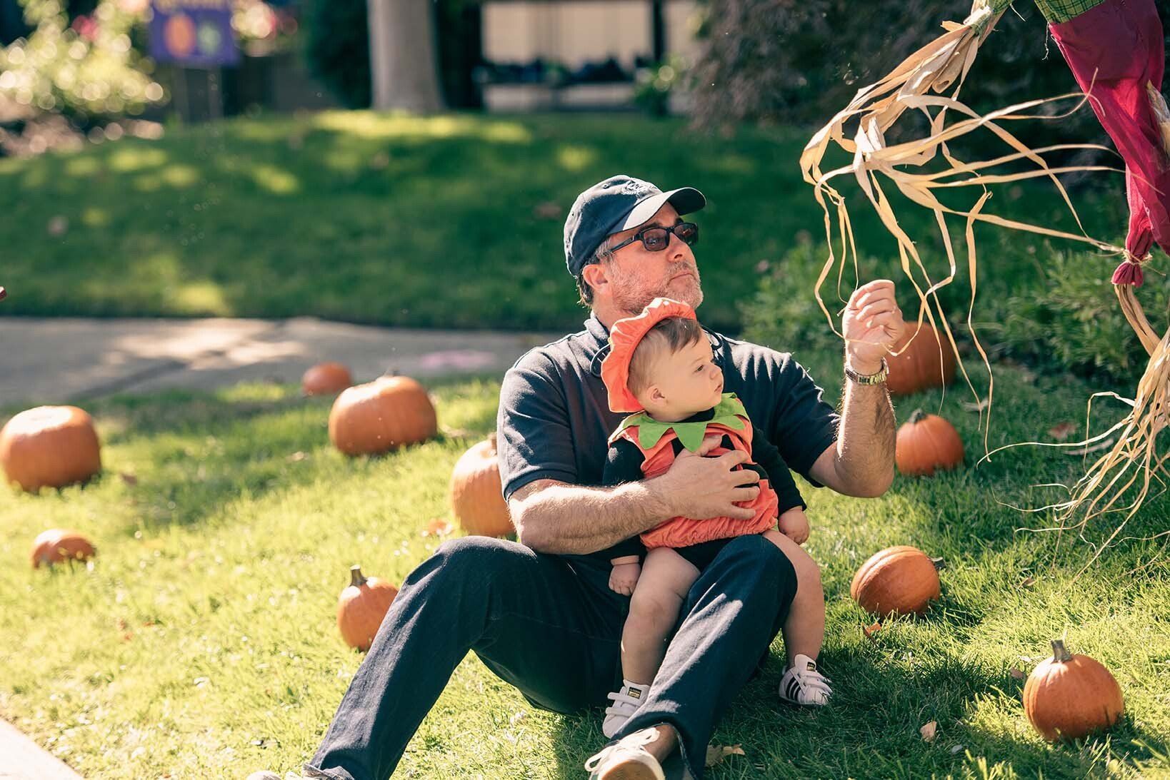 A man is sitting on the grass holding a baby in front of pumpkins.