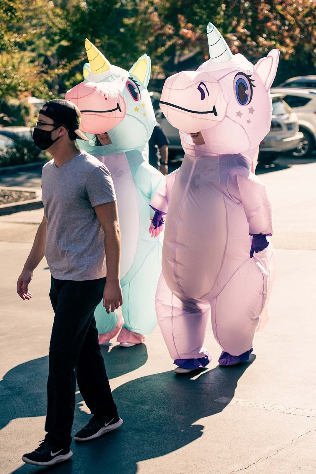 A man wearing a mask is walking next to two people in inflatable unicorn costumes.