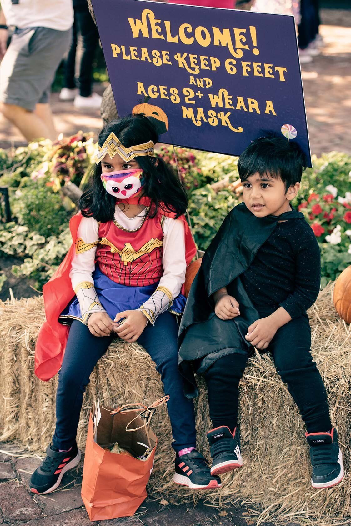 A boy and a girl in costumes are sitting on a hay bale holding a sign.