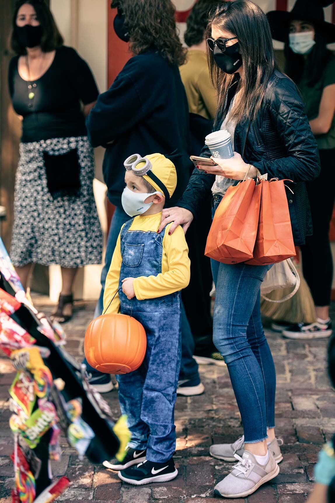 A woman is standing next to a child in a minion costume holding a pumpkin.