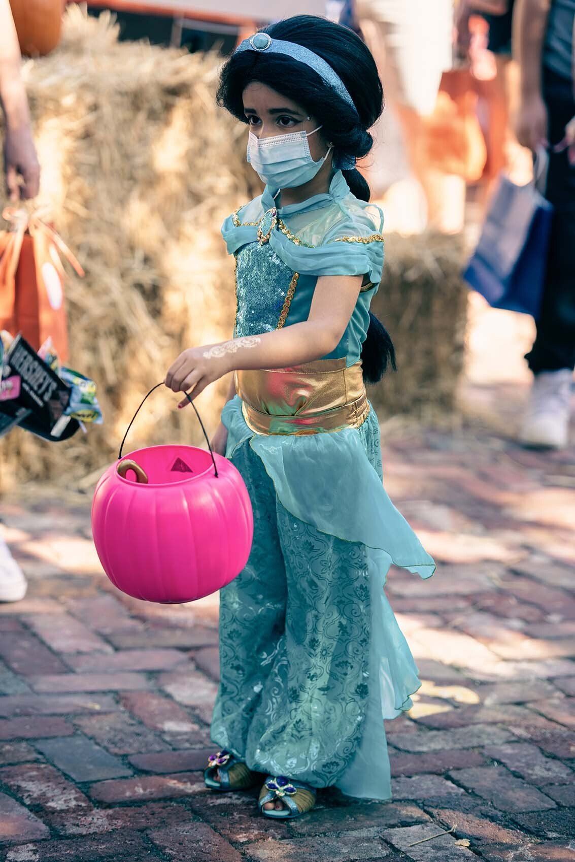 A little girl in a princess costume is wearing a mask and holding a pink pumpkin.