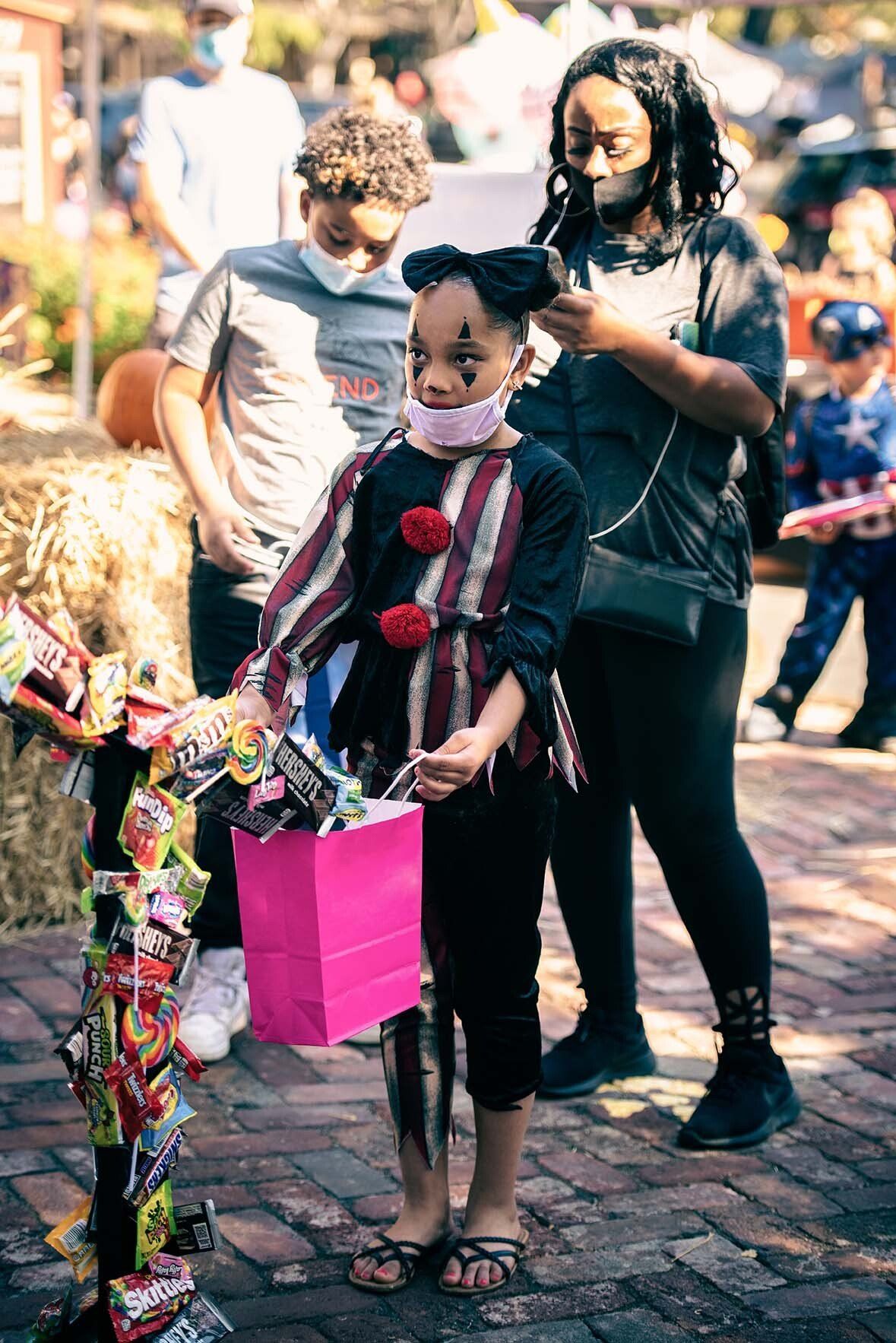 A little girl in a clown costume is holding a pink bag of candy.