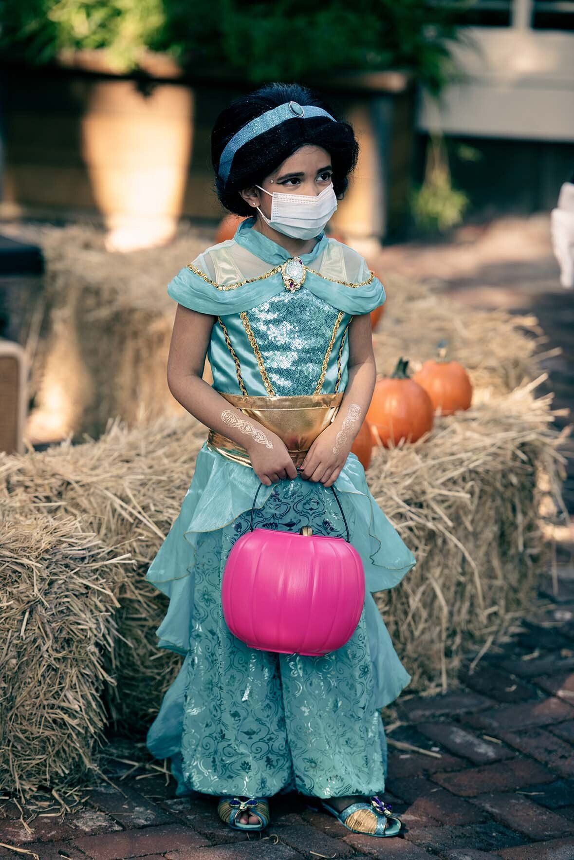 A little girl in a princess costume is wearing a mask and holding a pumpkin.