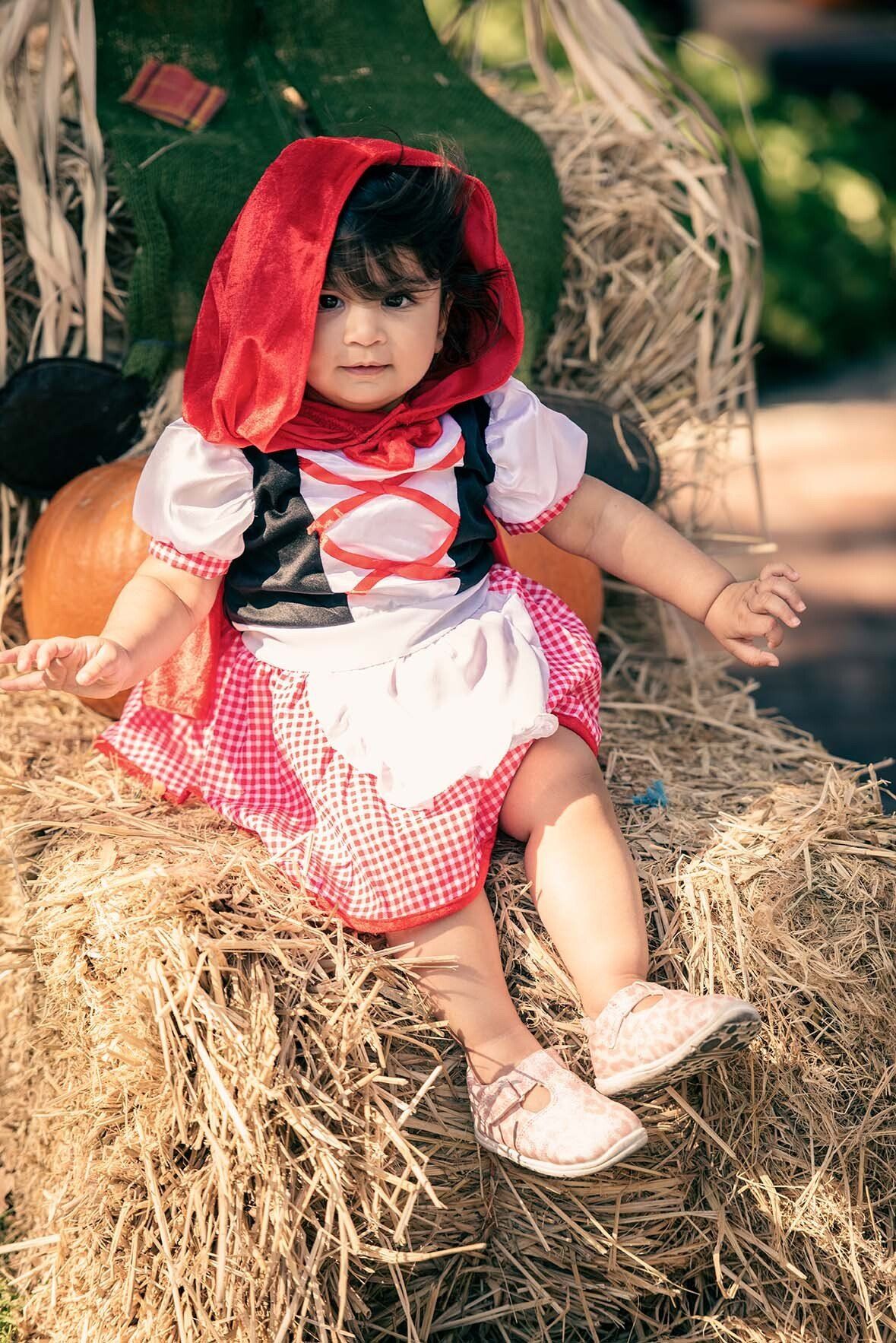 A little girl dressed as little red riding hood is sitting on a bale of hay.