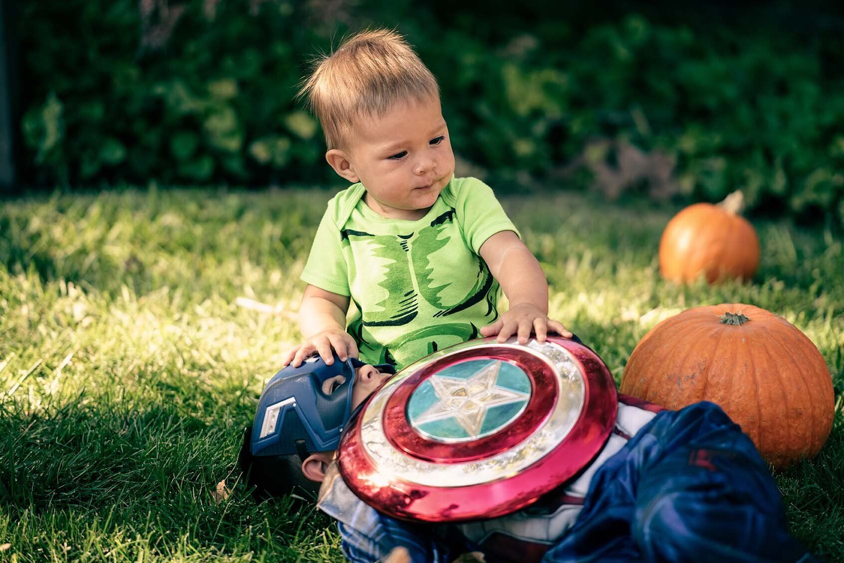 A little boy dressed as captain america is laying on top of a little boy dressed as hulk.