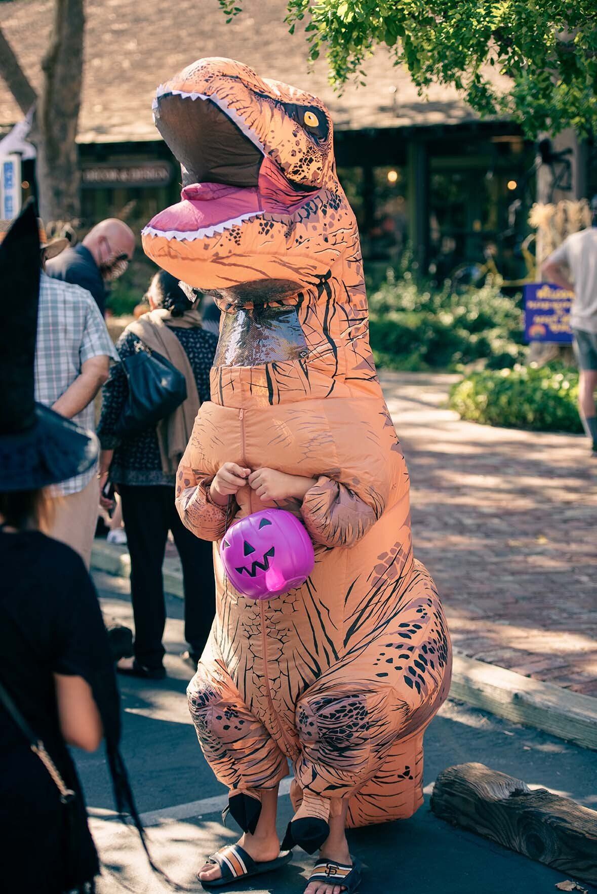 A person in a t-rex costume is holding a trick or treat bag.