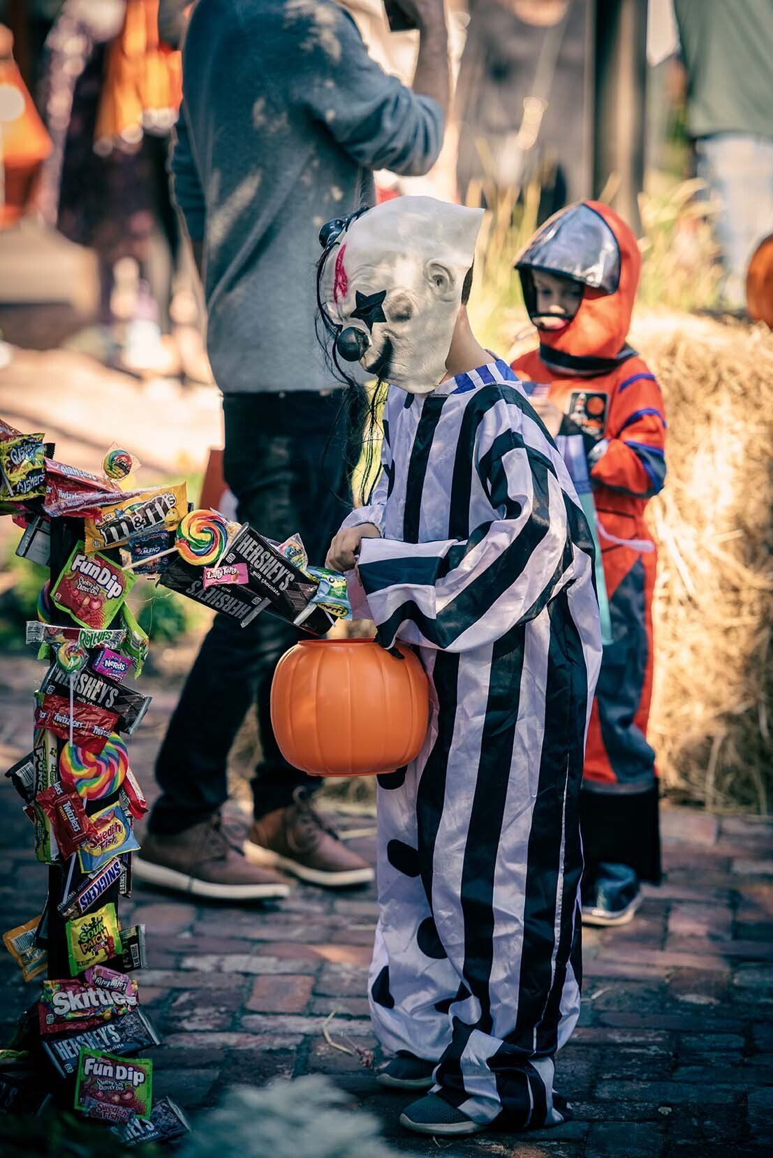 A child in a striped costume is holding a pumpkin and a bucket of candy.