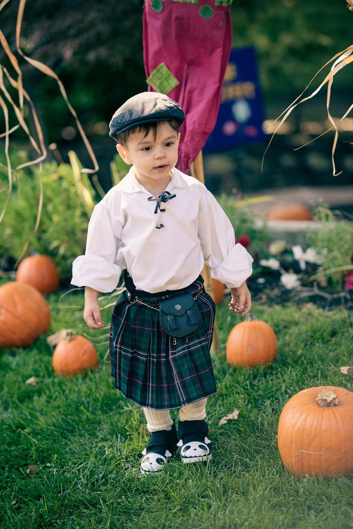 A little boy in a kilt is standing in the grass surrounded by pumpkins.