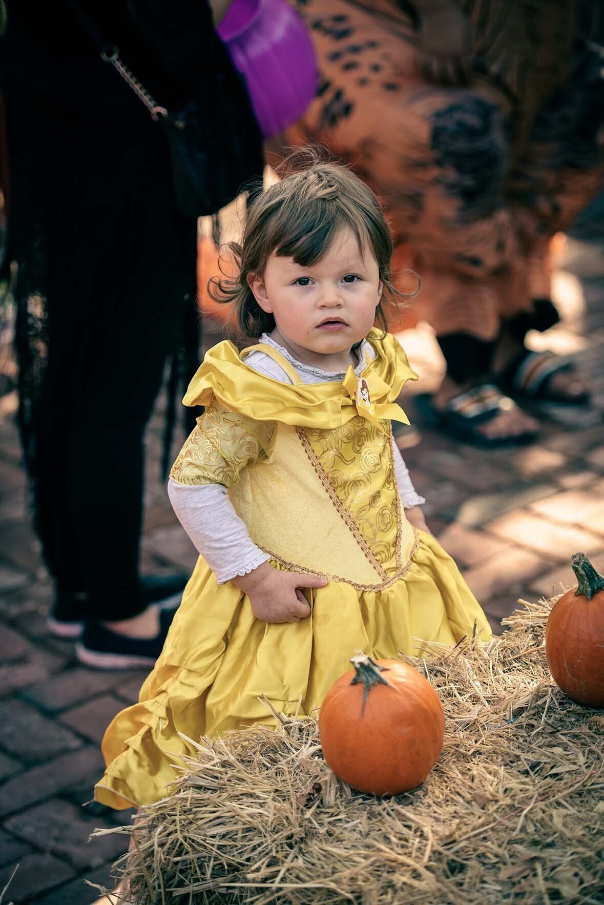 A little girl in a yellow dress is sitting on a bale of hay next to pumpkins.