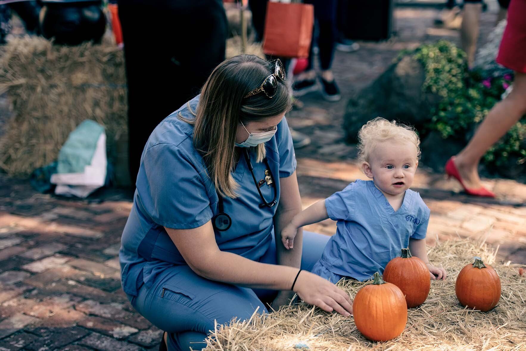 A nurse is kneeling down next to a baby playing with pumpkins.
