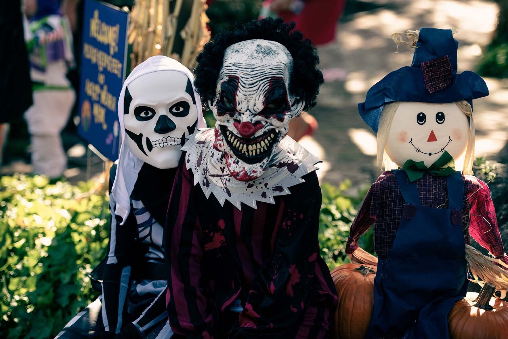 A scarecrow , a clown and a skeleton are sitting next to pumpkins.