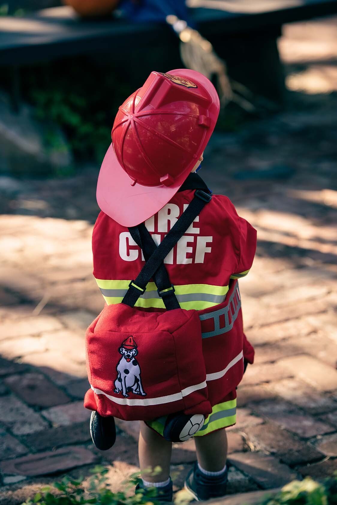A little boy in a fireman costume is standing on a brick sidewalk.