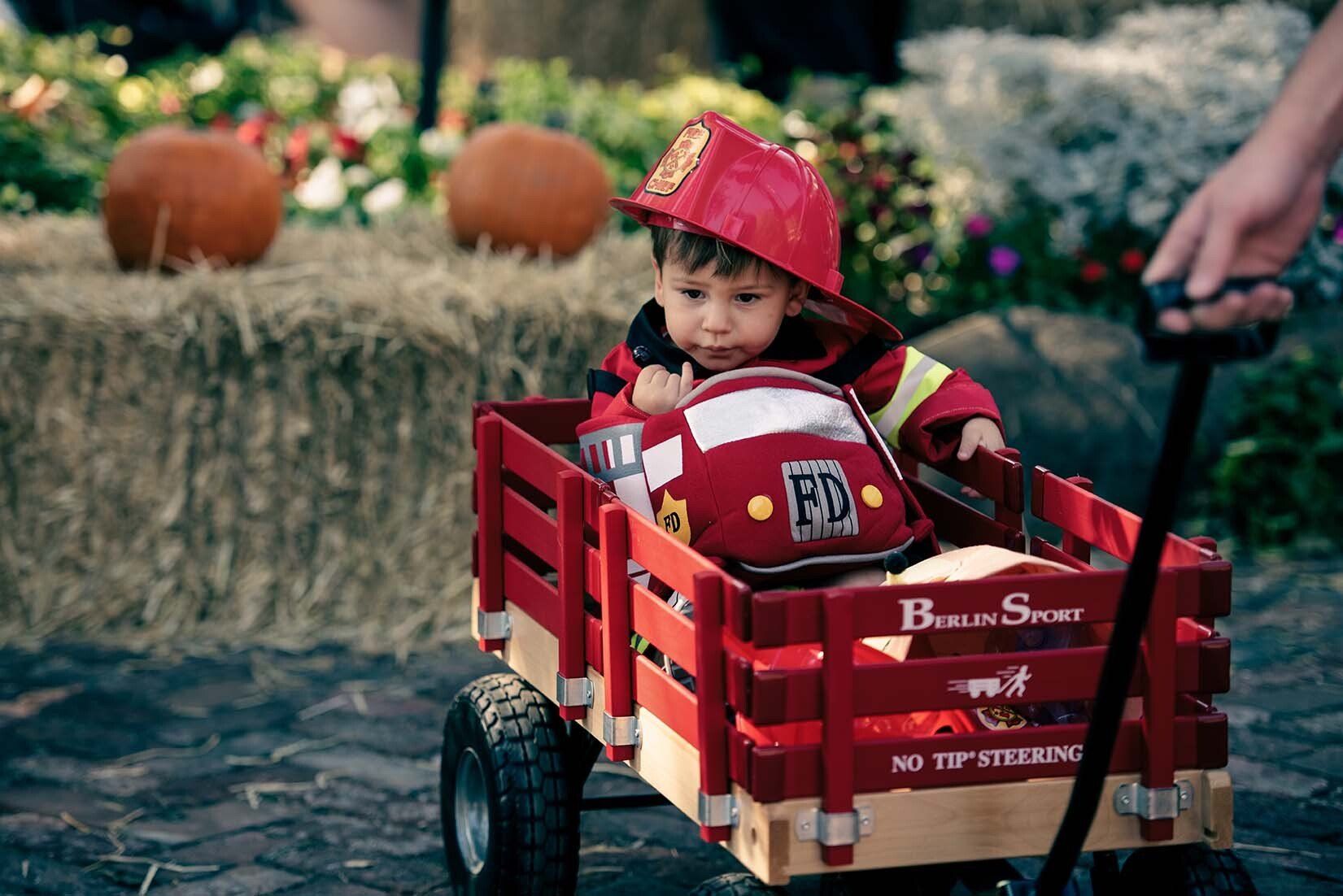 A baby in a fireman costume is sitting in a red wagon.