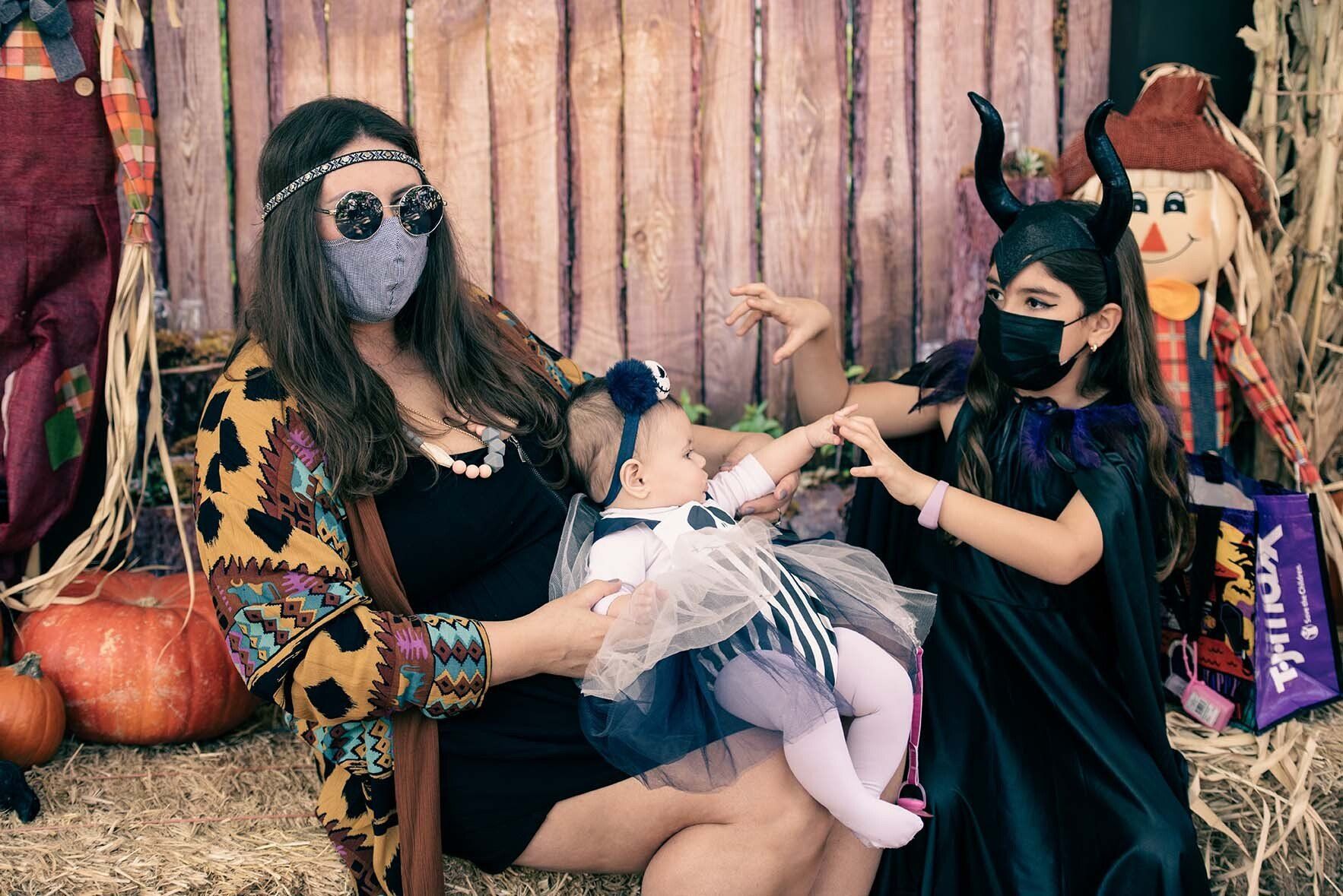 A woman wearing a mask is holding a baby and two girls in halloween costumes.