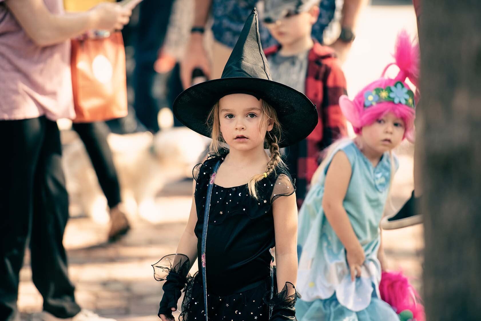 A little girl in a witch costume is standing in a crowd of people.