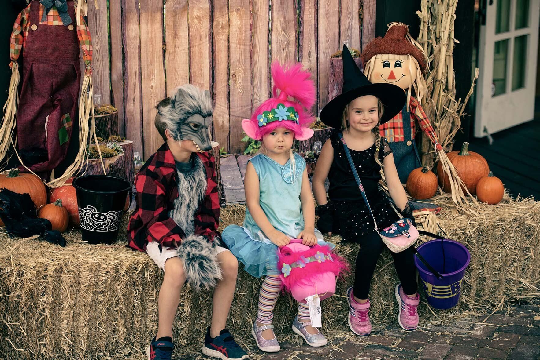 Three children in halloween costumes are sitting on hay bales.