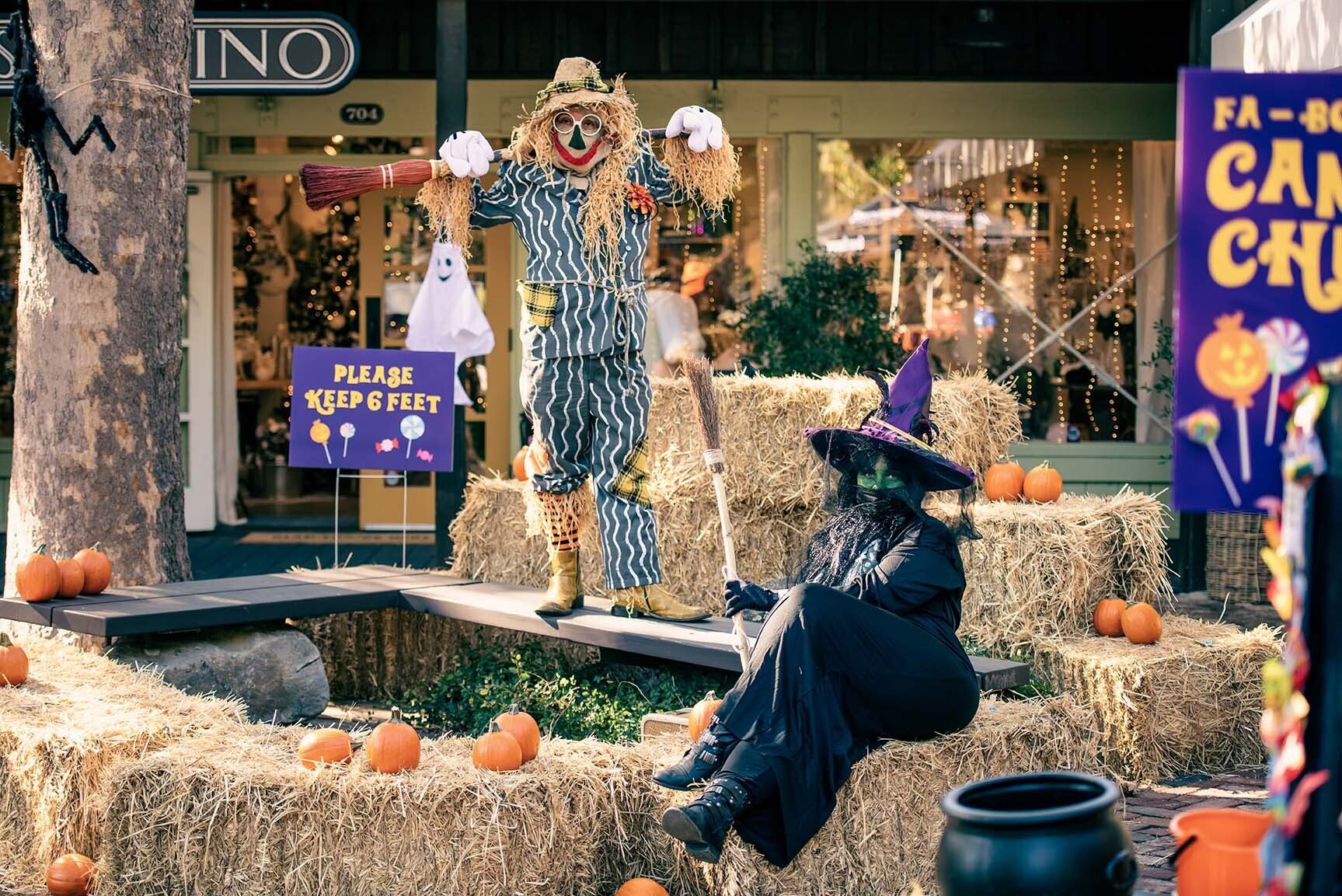 A scarecrow and a witch are sitting on hay bales in front of a store.