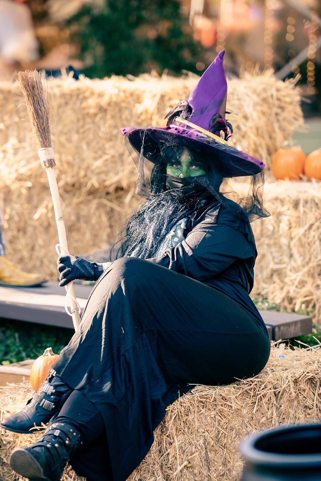 A woman dressed as a witch is sitting on a bale of hay holding a broom.
