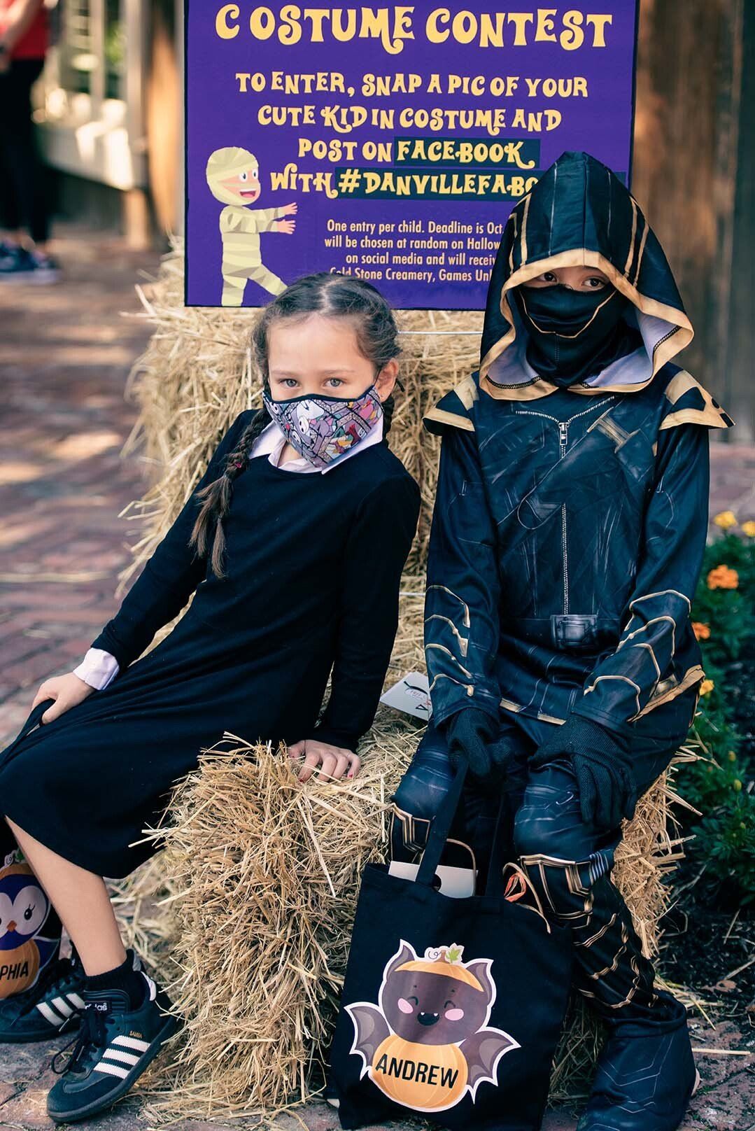 Two children in costumes are sitting on a hay bale.