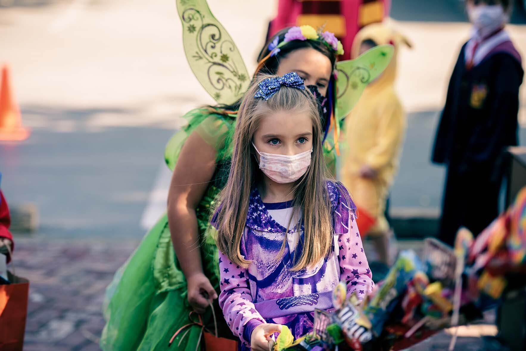 A girl wearing a mask and a fairy costume is holding a trick or treat bag.