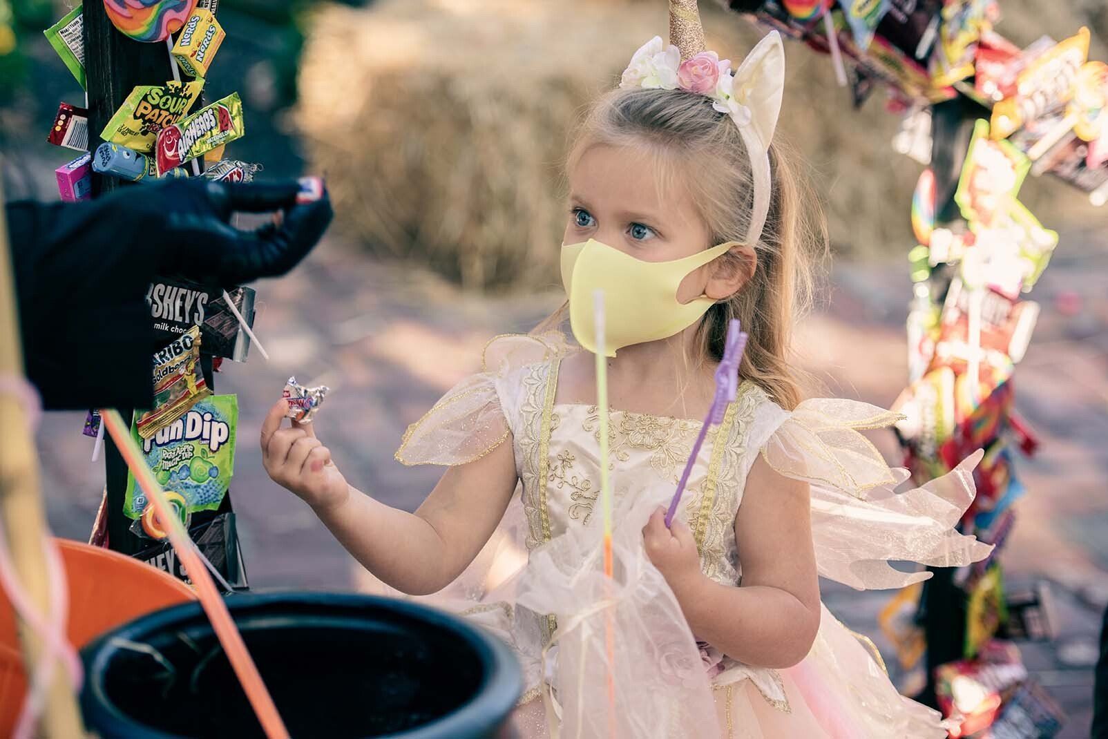 A little girl wearing a mask and a fairy costume is holding a lollipop.