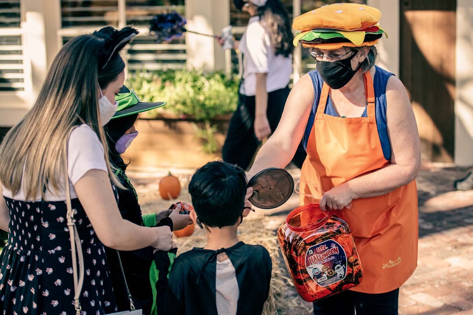 A woman in a hamburger hat is handing out candy to children at a trick or treat event.