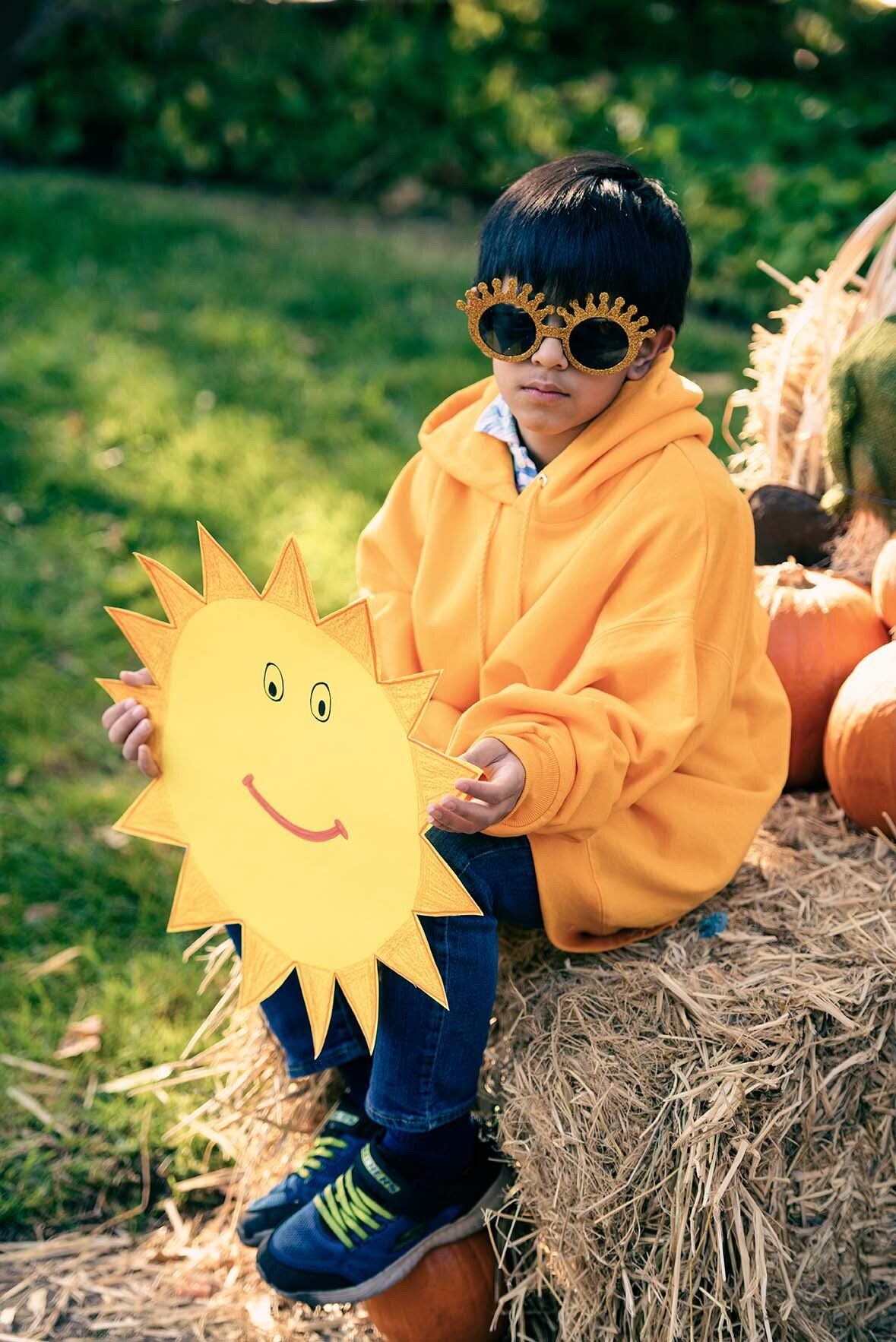 A young boy is sitting on a bale of hay holding a paper sun.