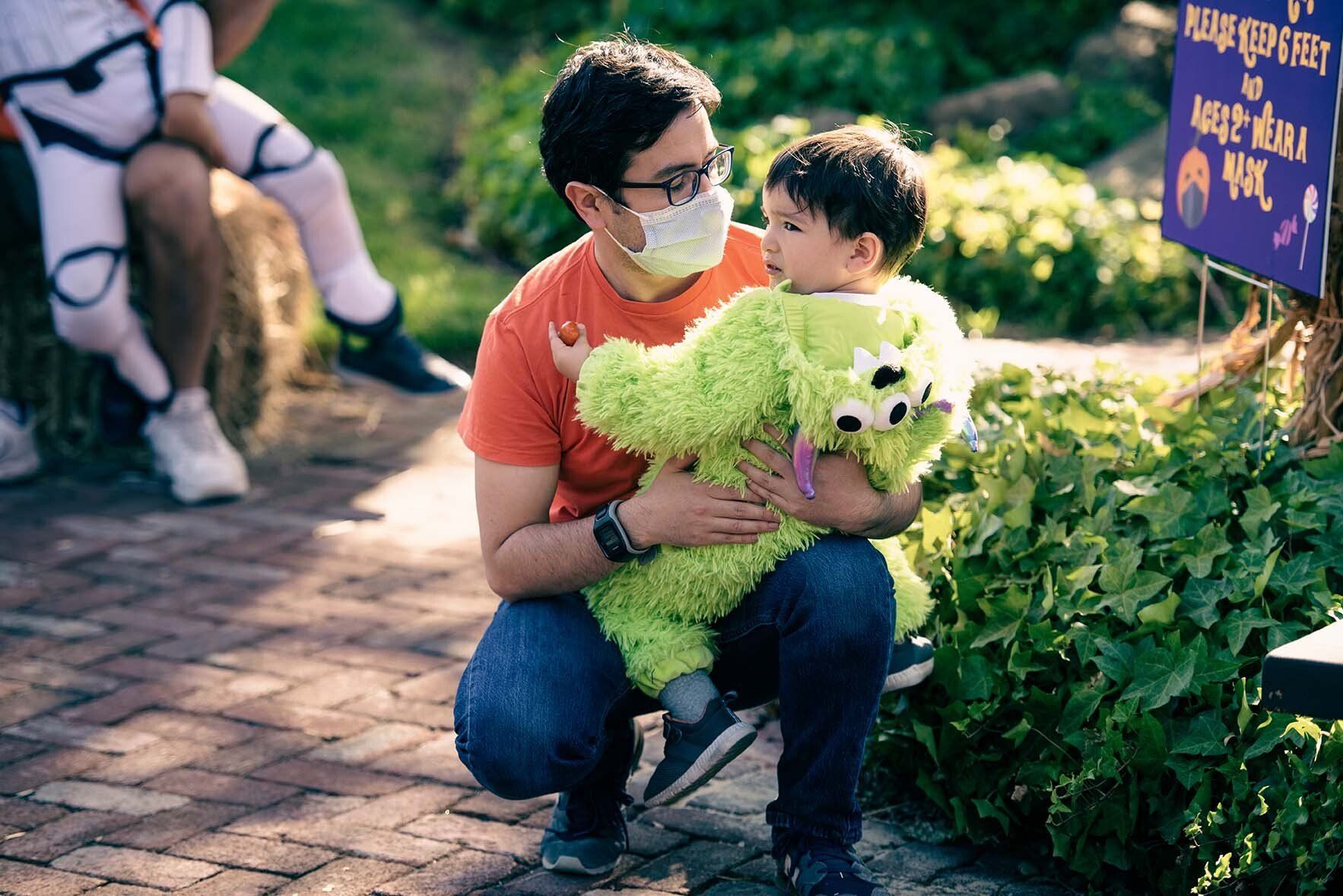 A man wearing a mask is holding a baby in a green costume.