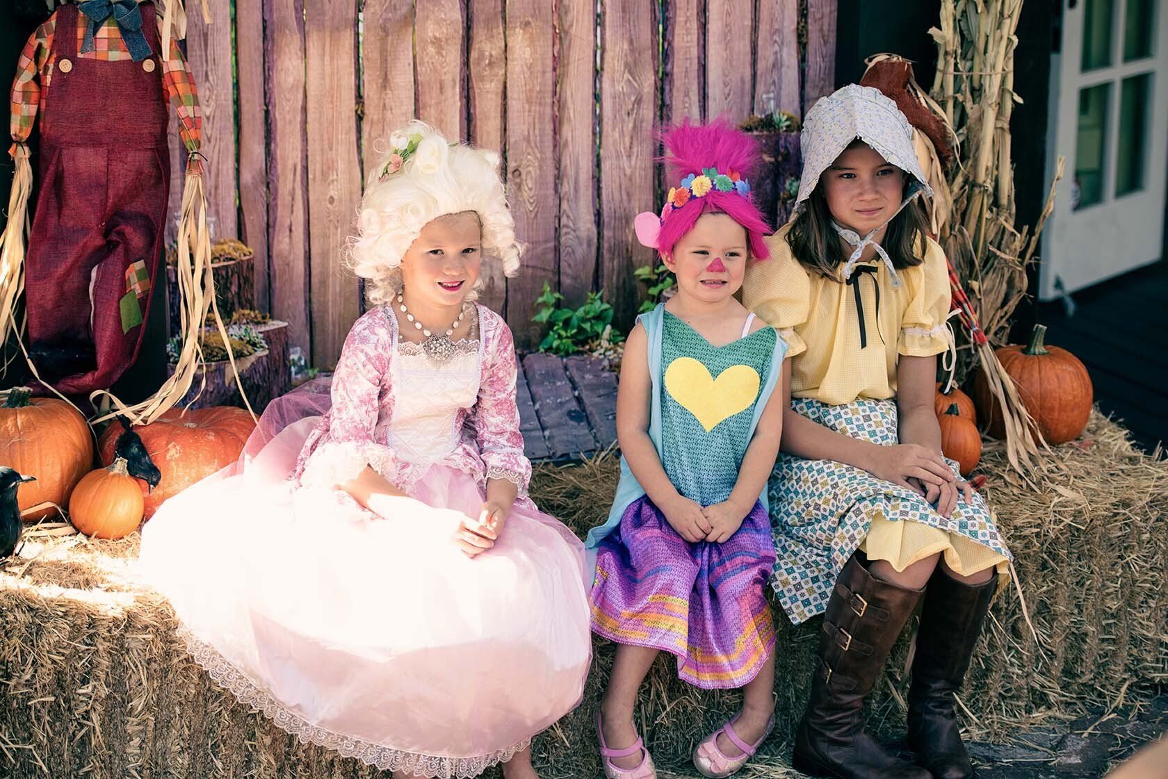 Three little girls in costumes are sitting on a bale of hay.