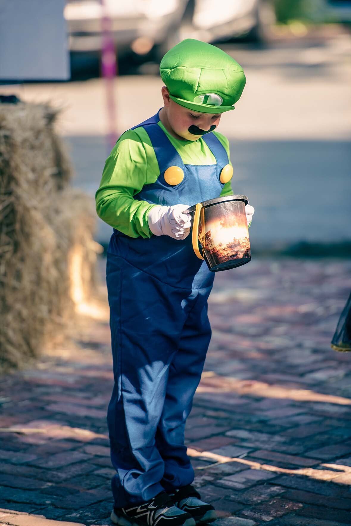 A young boy in a luigi costume is holding a bucket of treats.