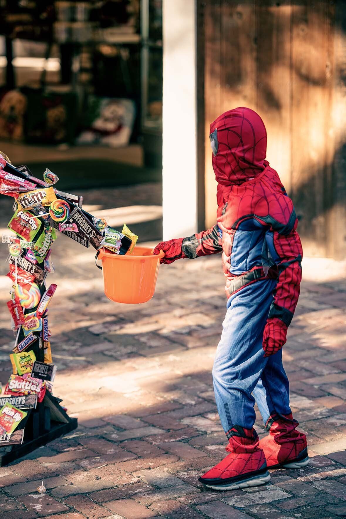 A young boy in a spiderman costume is holding a trick or treat bucket.