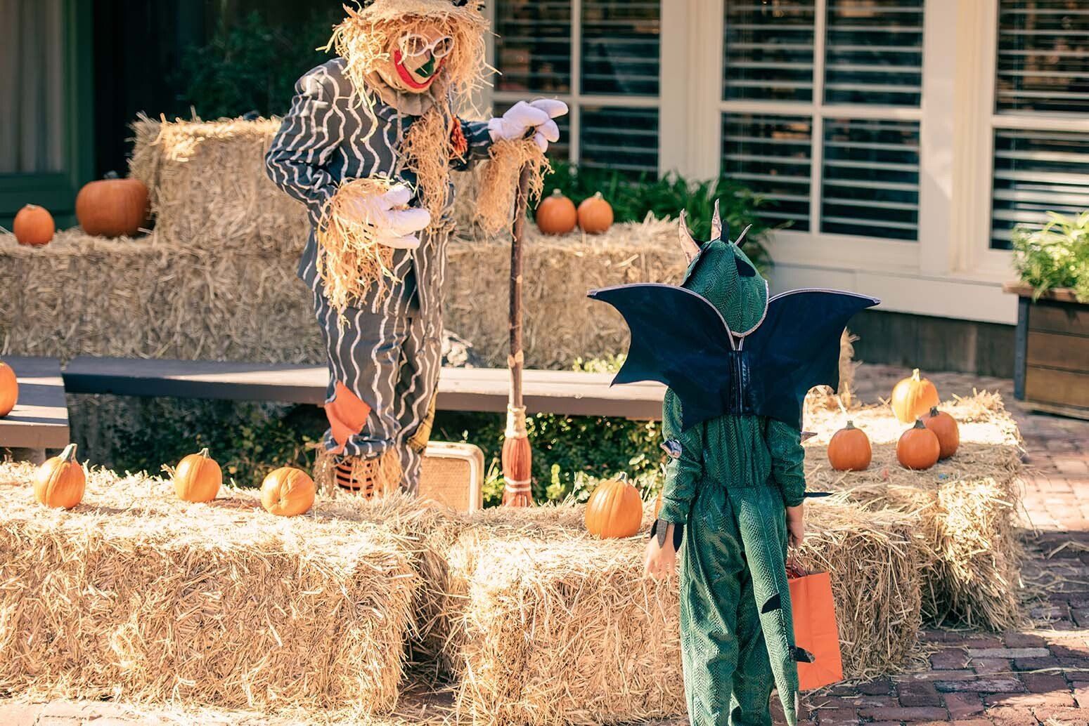 A scarecrow and a child in a halloween costume are standing next to hay bales.