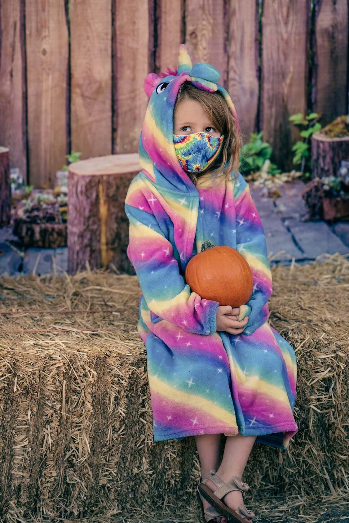 A little girl in a unicorn costume is sitting on a bale of hay holding a pumpkin.
