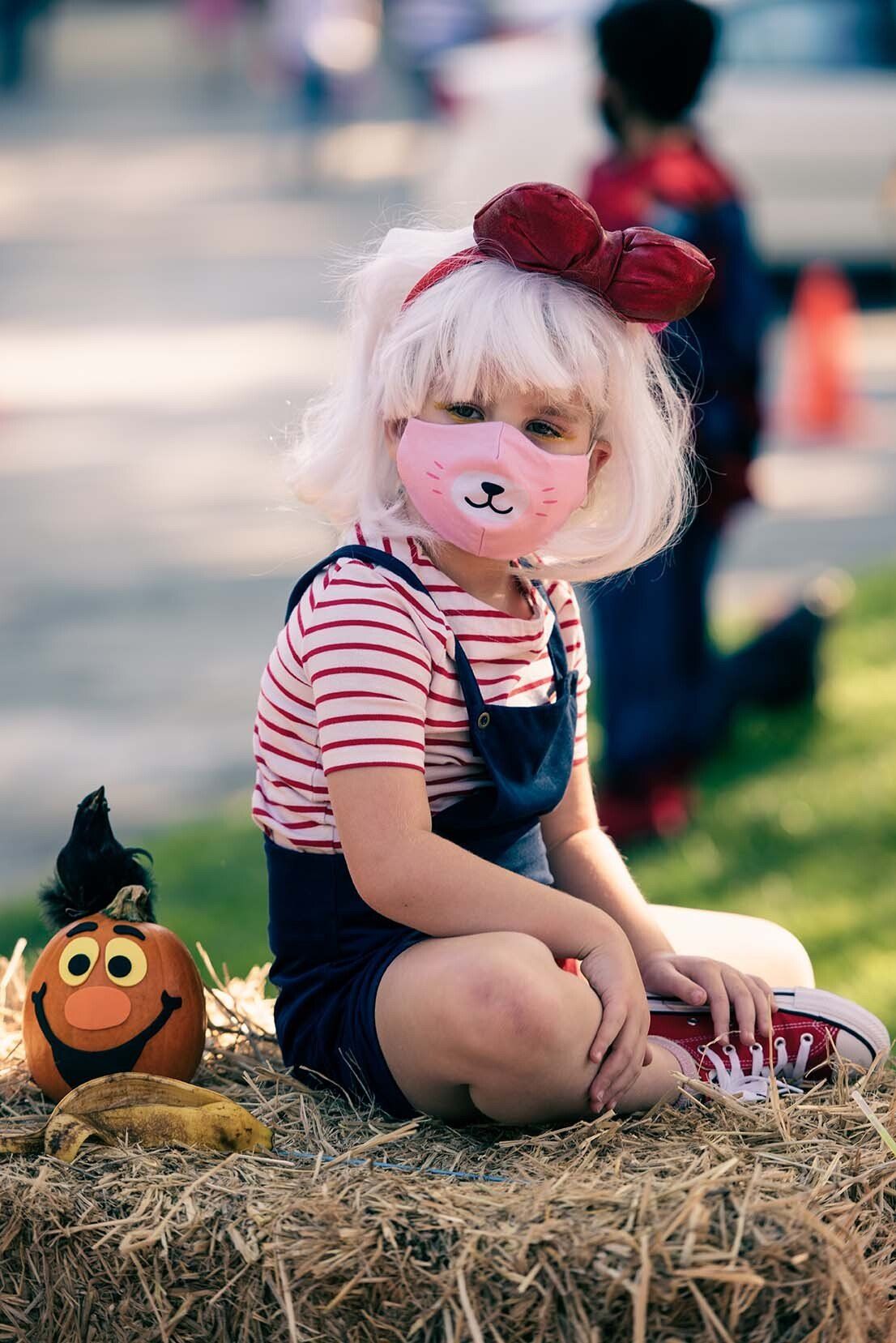 A little girl wearing a mask is sitting on a bale of hay.