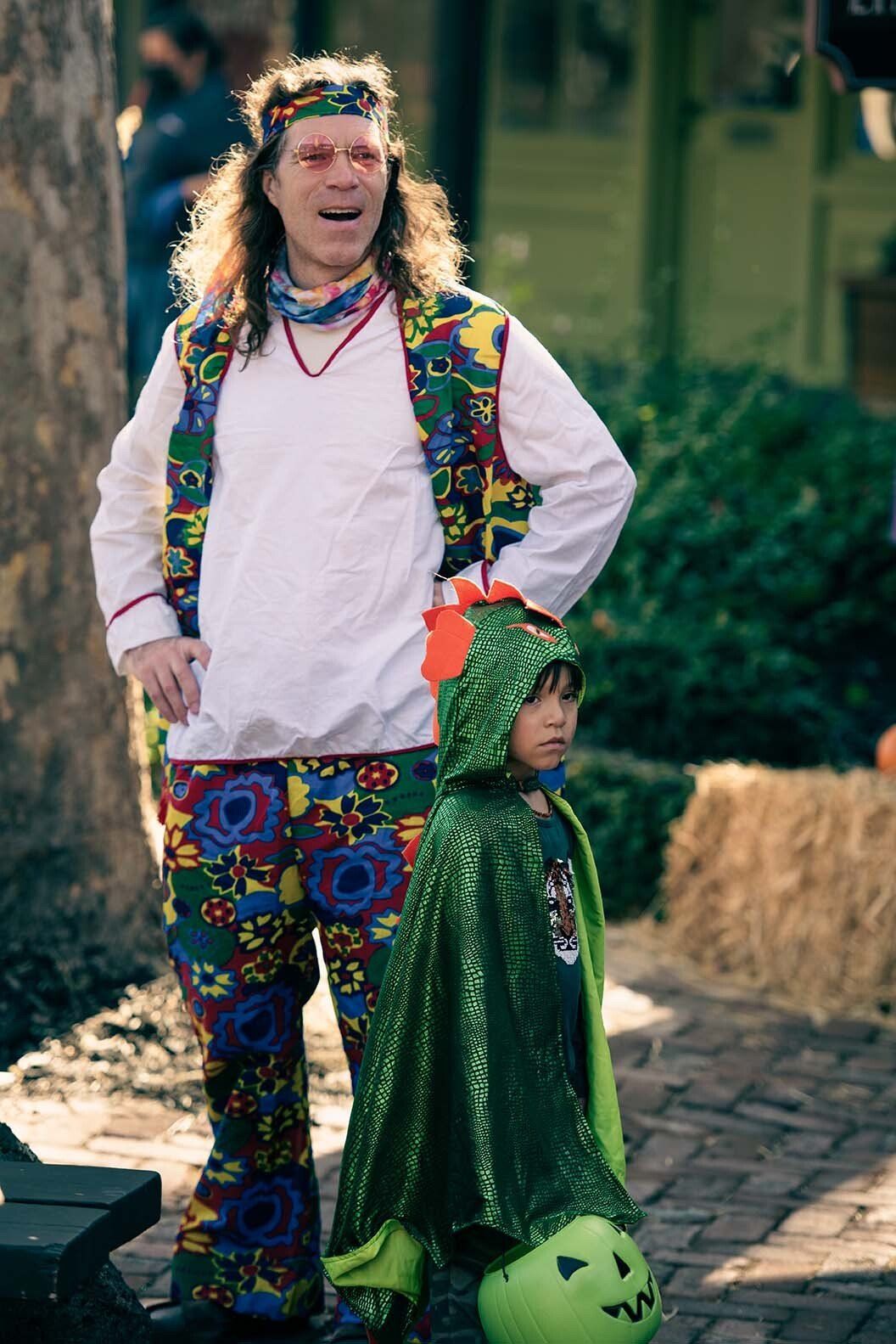 A man in a hippie costume is standing next to a child in a green cape.