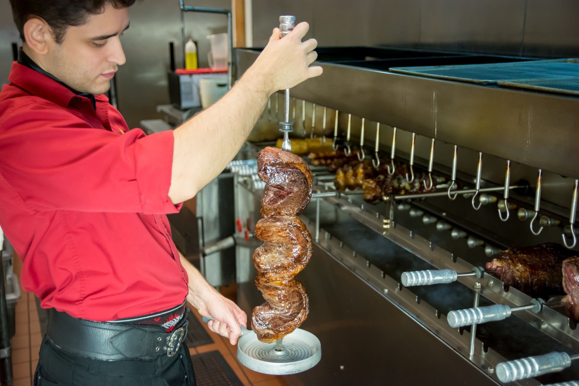 A man in a red shirt is cutting a piece of meat on a skewer.