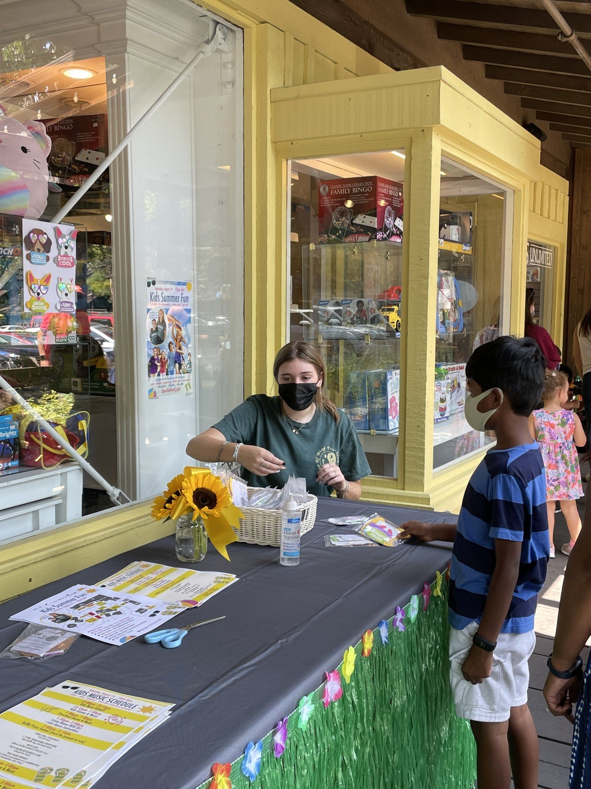 A woman wearing a mask is talking to a boy at a table in front of a store.