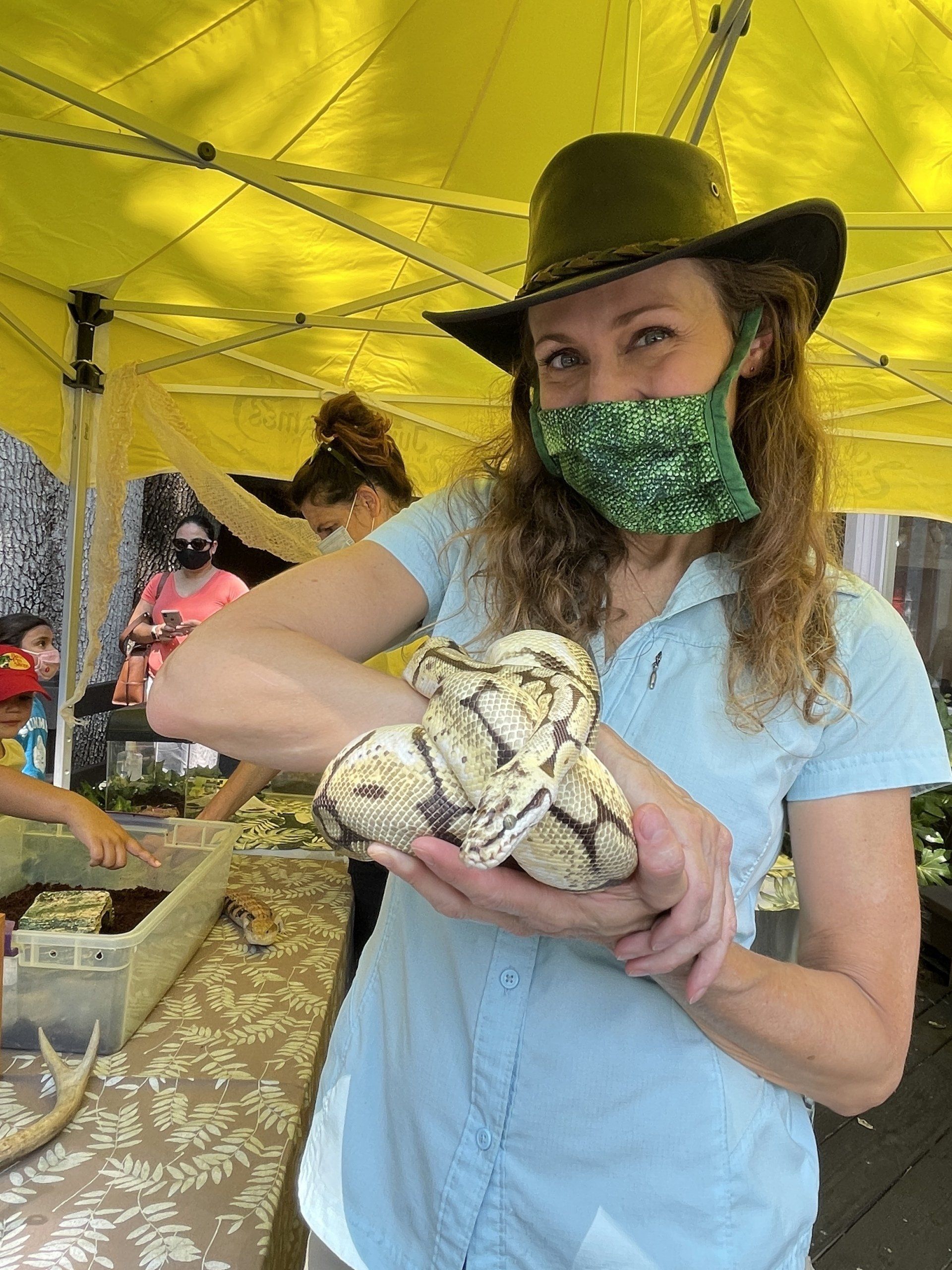 A woman wearing a cowboy hat and a mask is holding a snake.