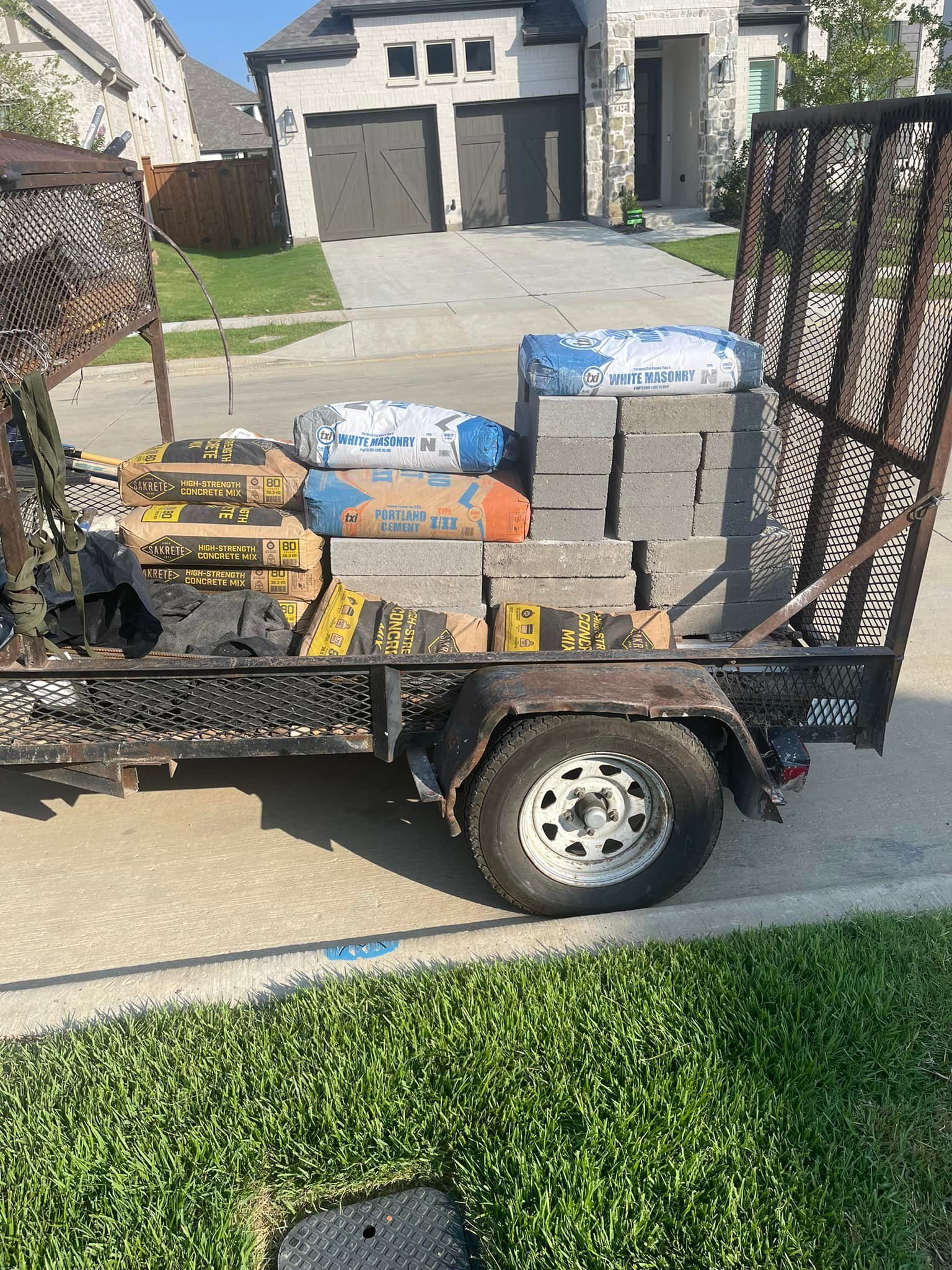 A trailer filled with bricks and bags of cement is parked in front of a house.