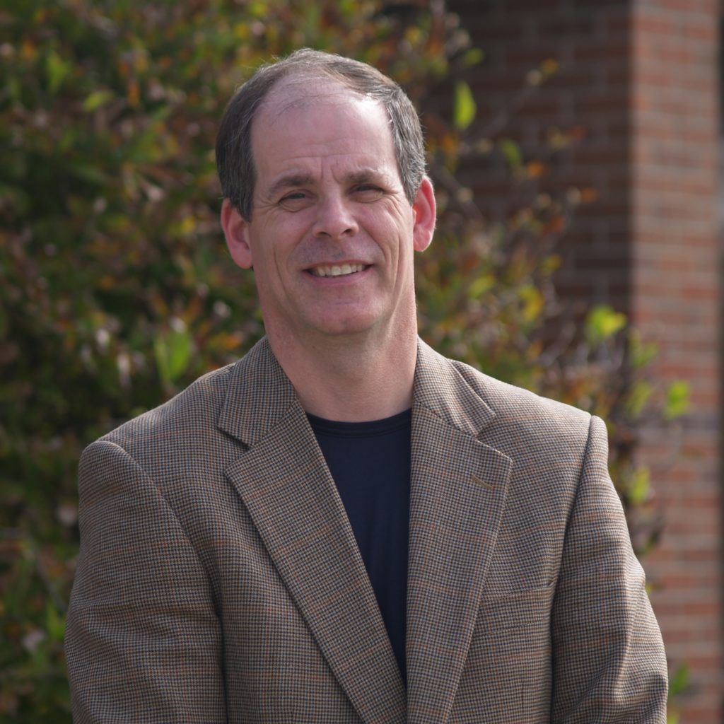Man in blazer smiles outdoors near brick building and greenery.