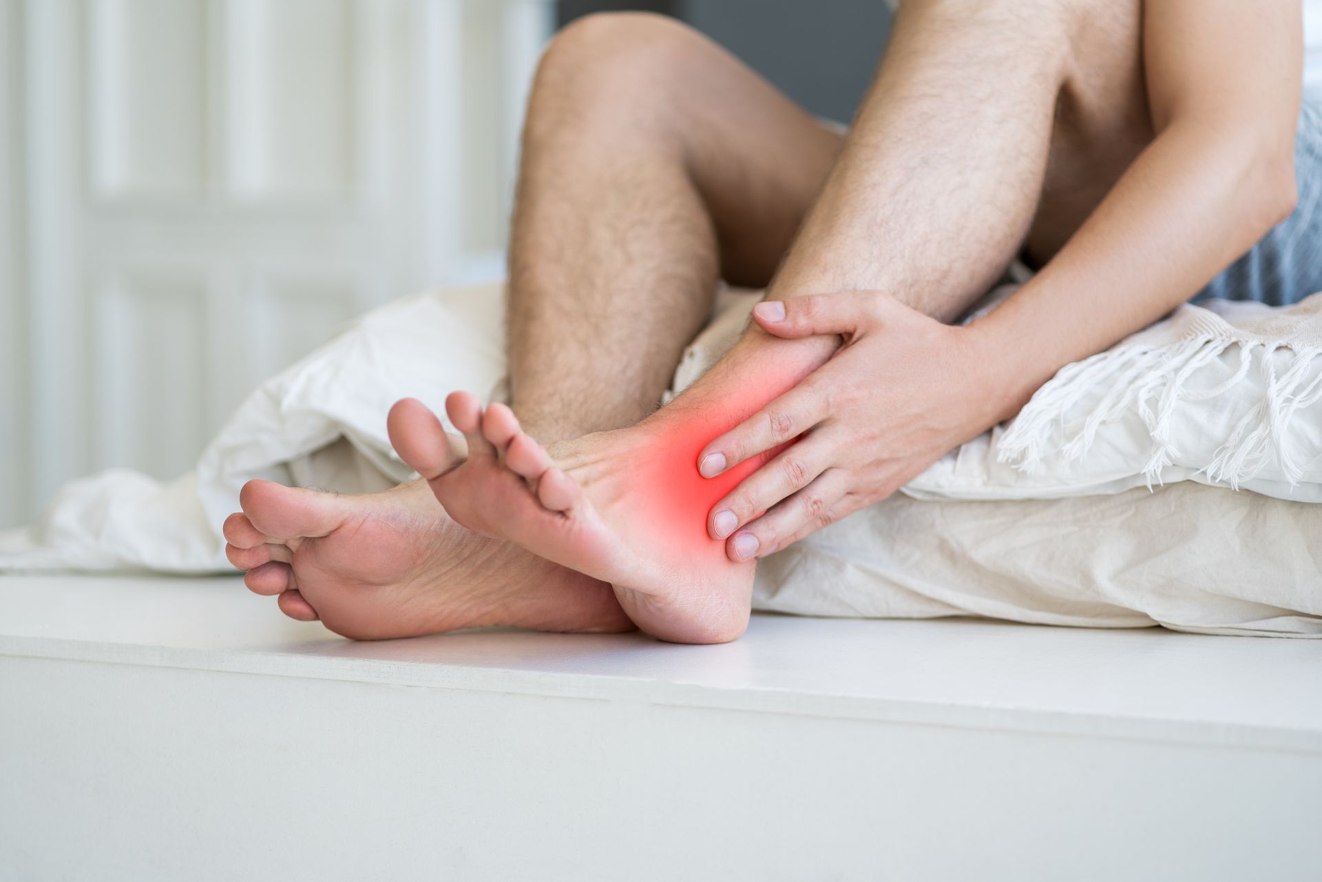 Person sitting on a bed, holding their foot with a red-highlighted ankle, indicating pain.