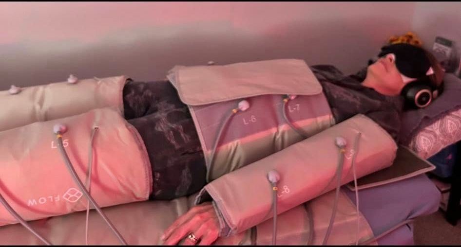 Person lying in a wellness device, eyes covered, wearing headphones. They are surrounded by padded sleeves and tubes in a room with red lighting.