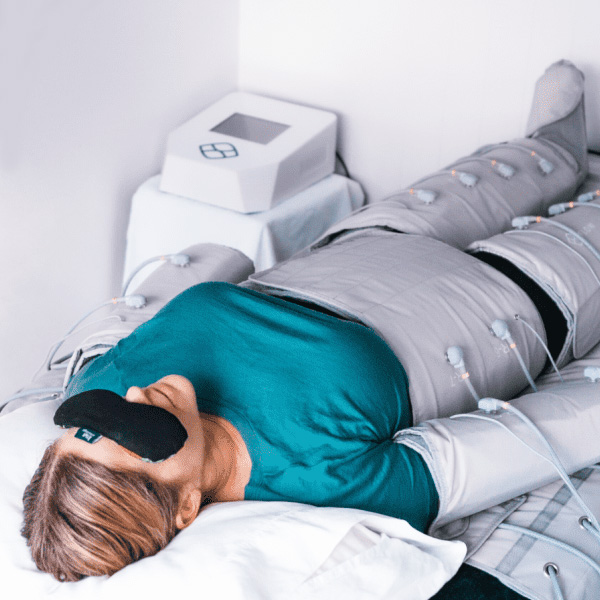 Woman lying down, wearing a compression therapy suit. She has a black eye mask on, and the machine is in a white room.