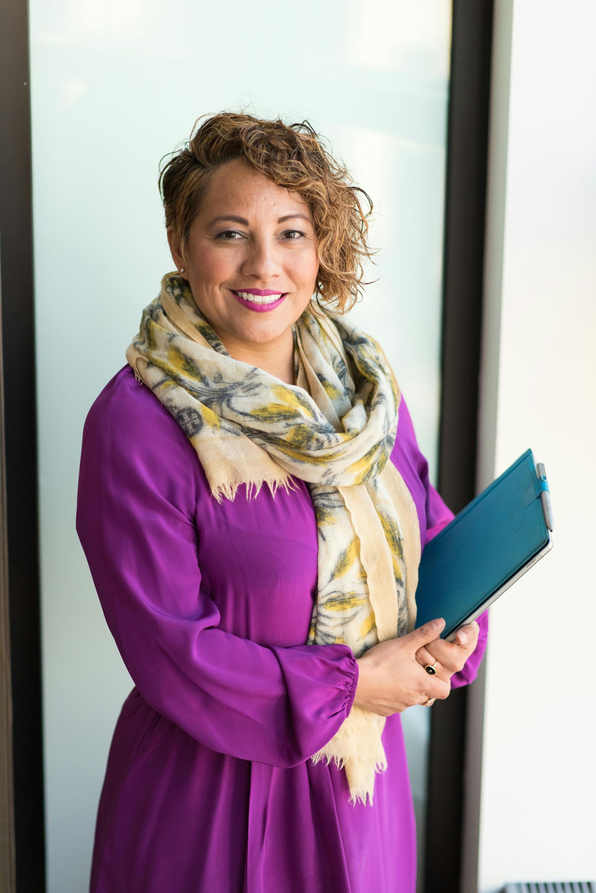 Woman with curly brown hair, wearing a purple dress and patterned scarf, holding a blue notebook.