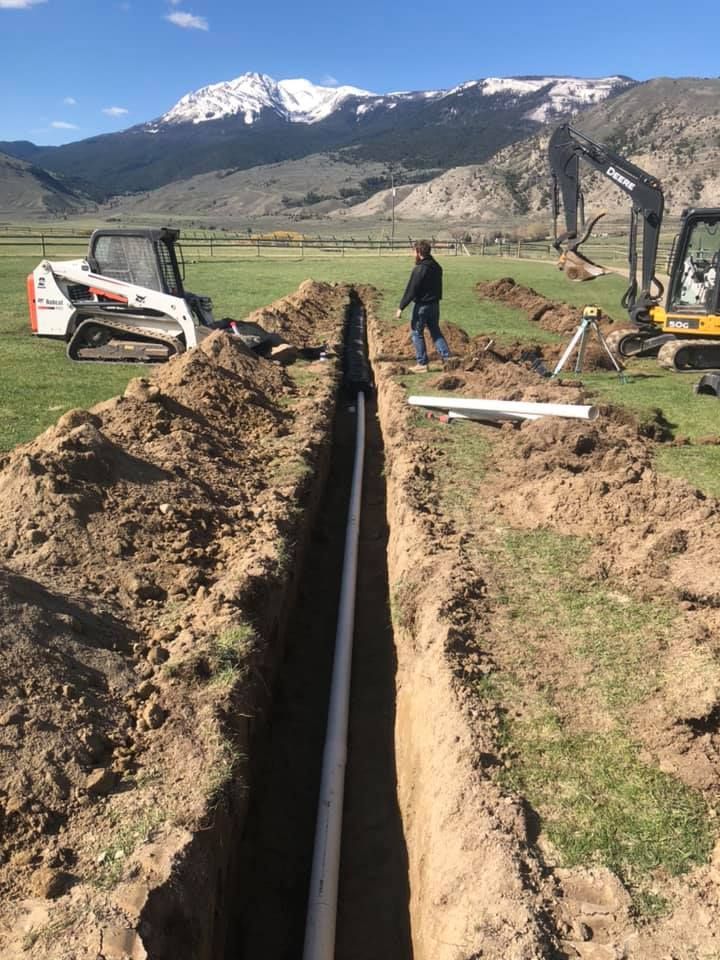 Trench dug in a field with white PVC pipe, two excavators, and snow-capped mountains in the background.
