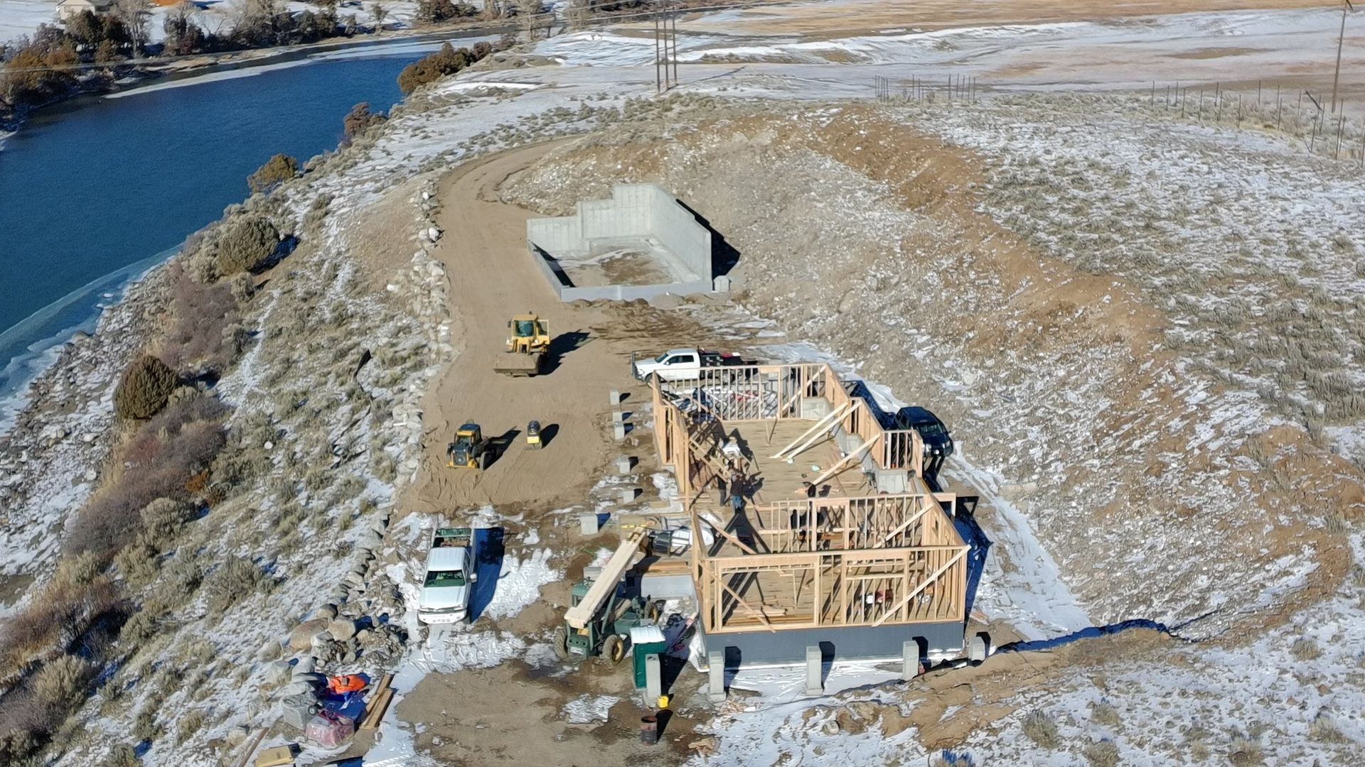 Construction site with partially built house and foundation, overlooking a river. Snowy hillside.