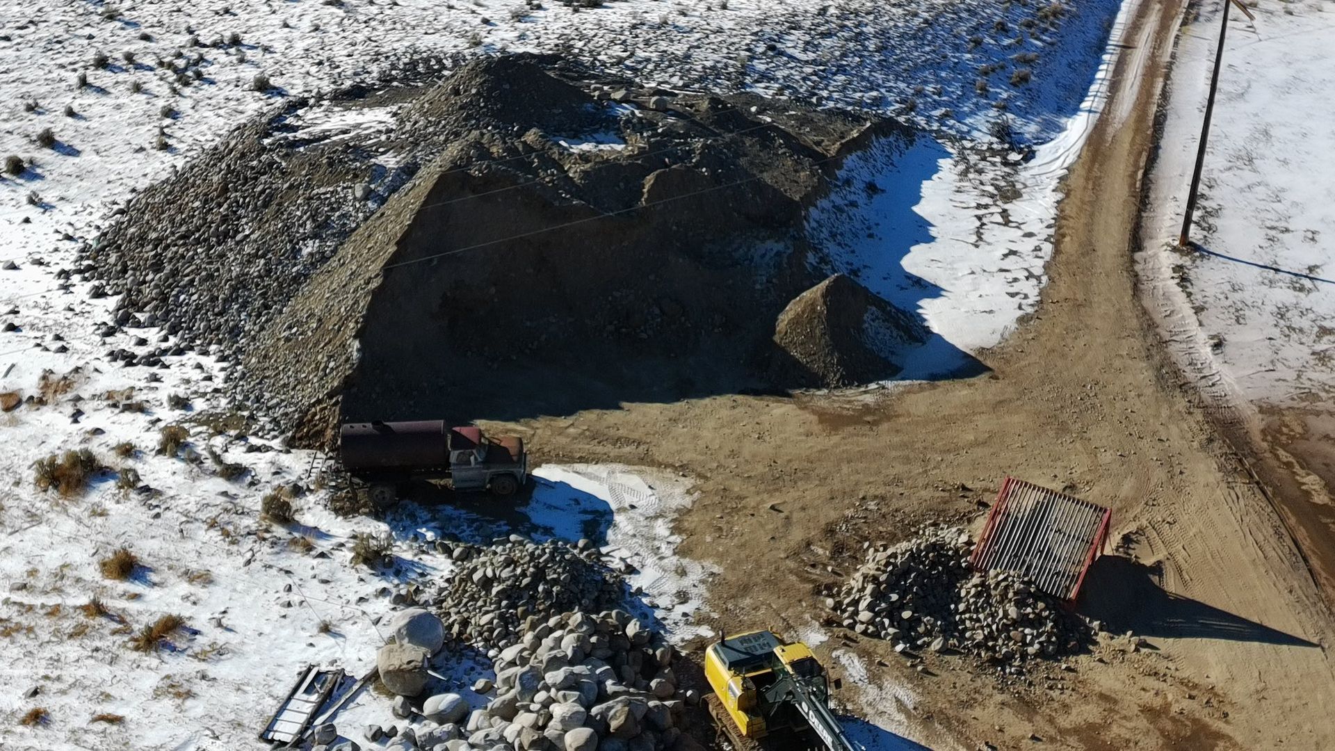An aerial view of a gravel pile, with two trucks and an excavator on a snowy landscape.